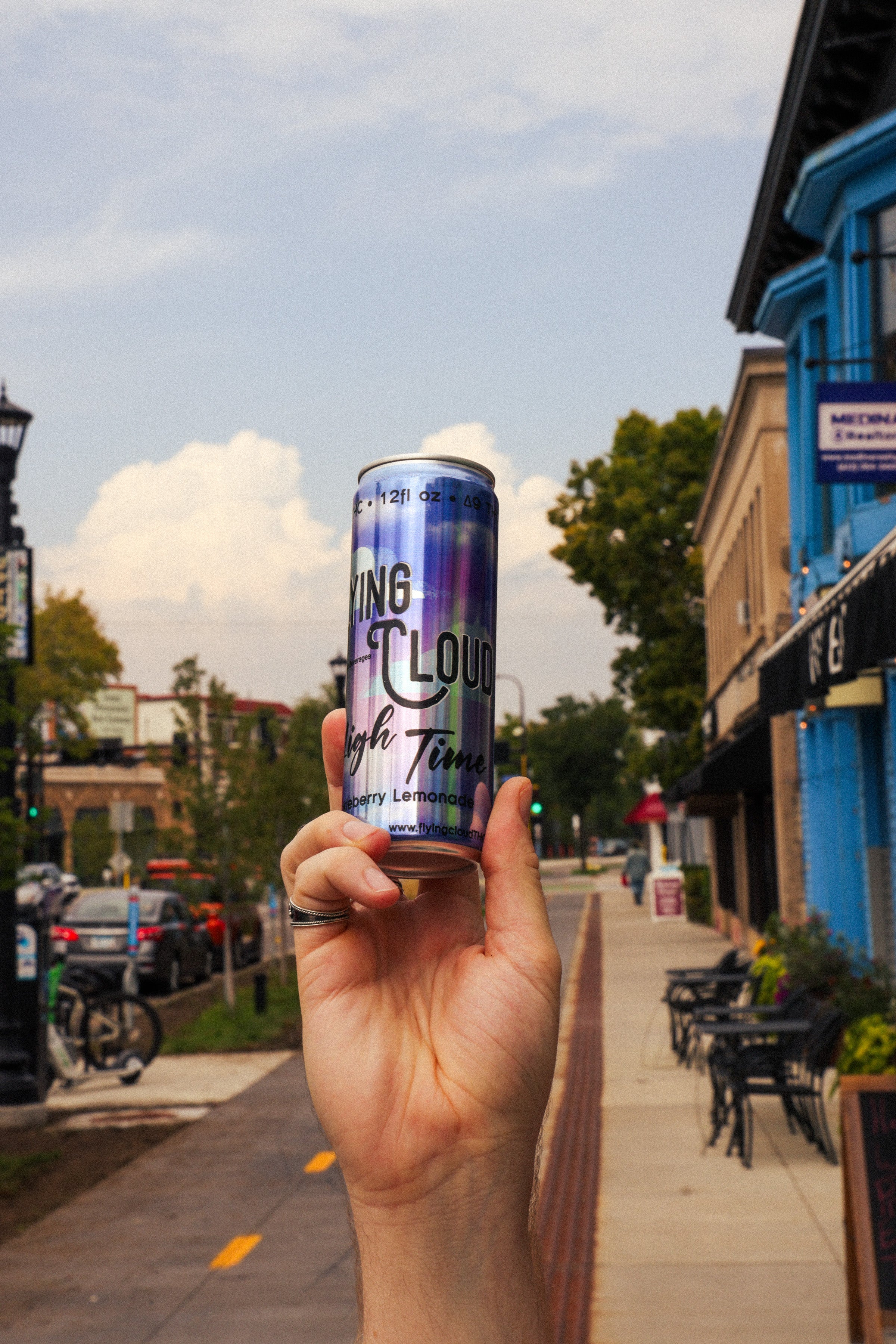 Hand holding a Flying Cloud THC drink can with a city street background